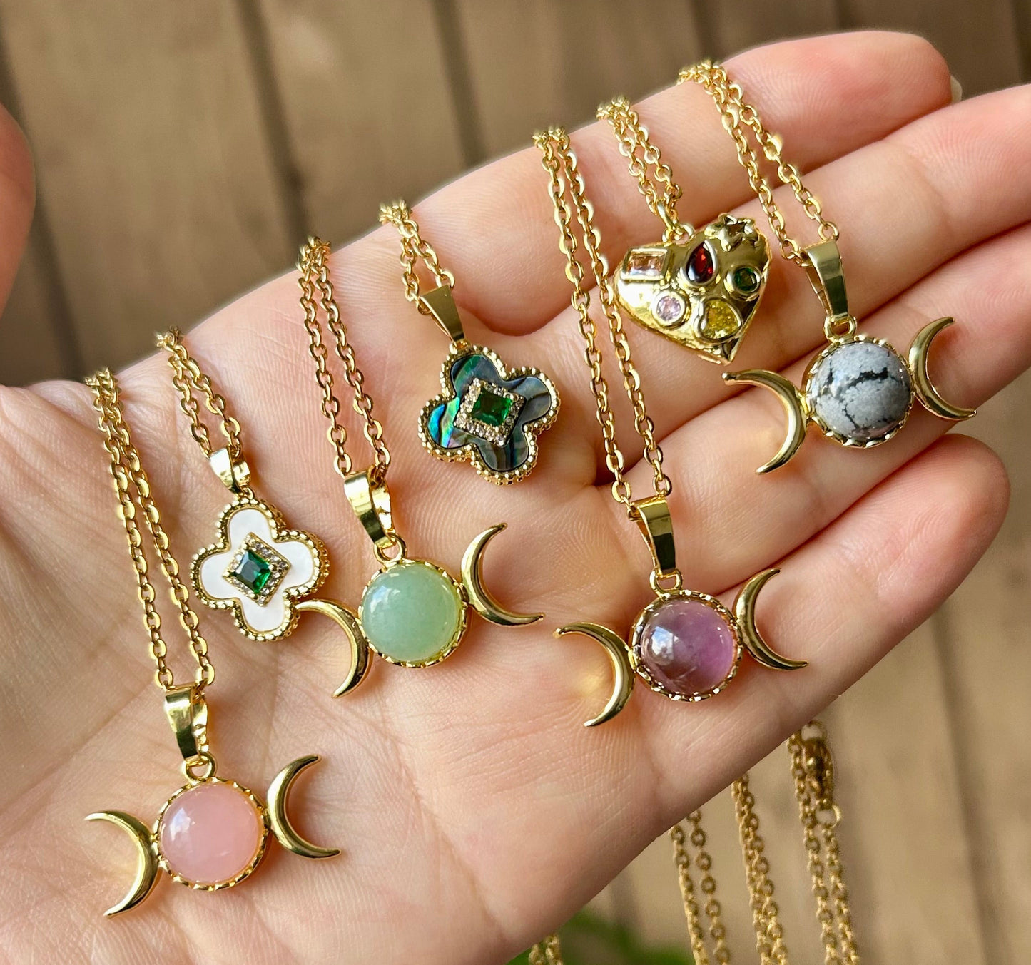 Hand holding multiple gold necklaces with various pendants against a blurred natural background