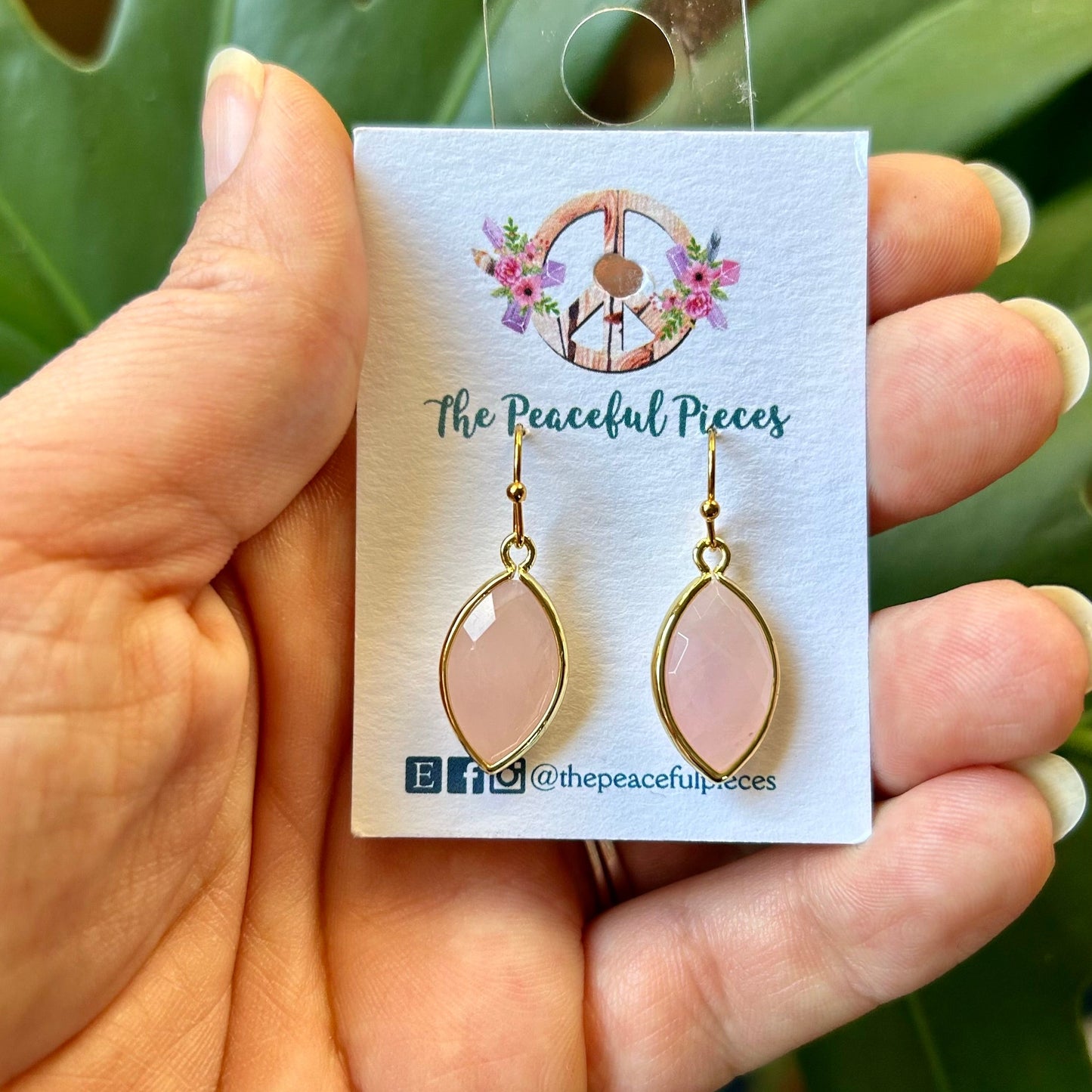 Pink earrings on a 'The Peaceful Pieces' card held by a hand with green leaves in the background