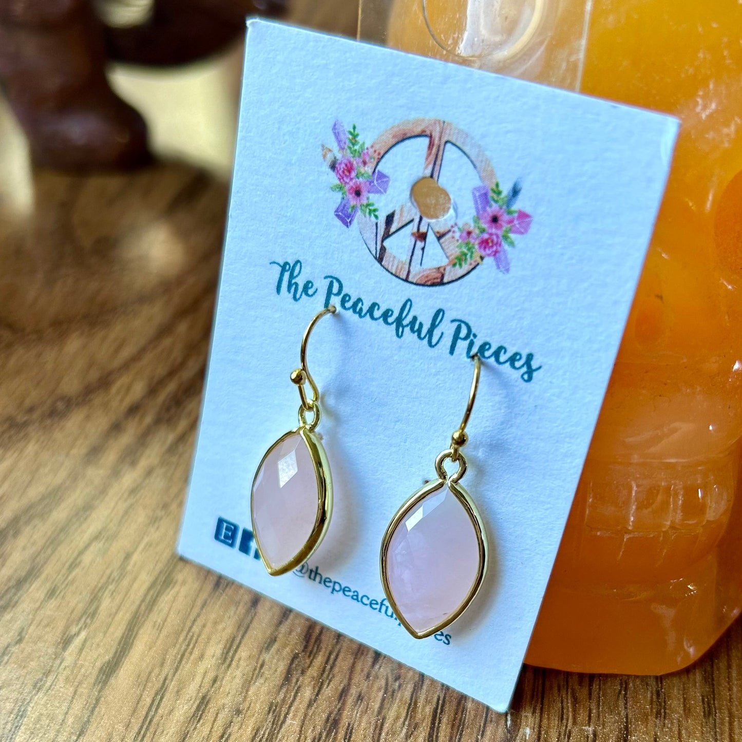 Gold earrings on a card with 'The Peaceful Pieces' branding, displayed on a wooden surface.