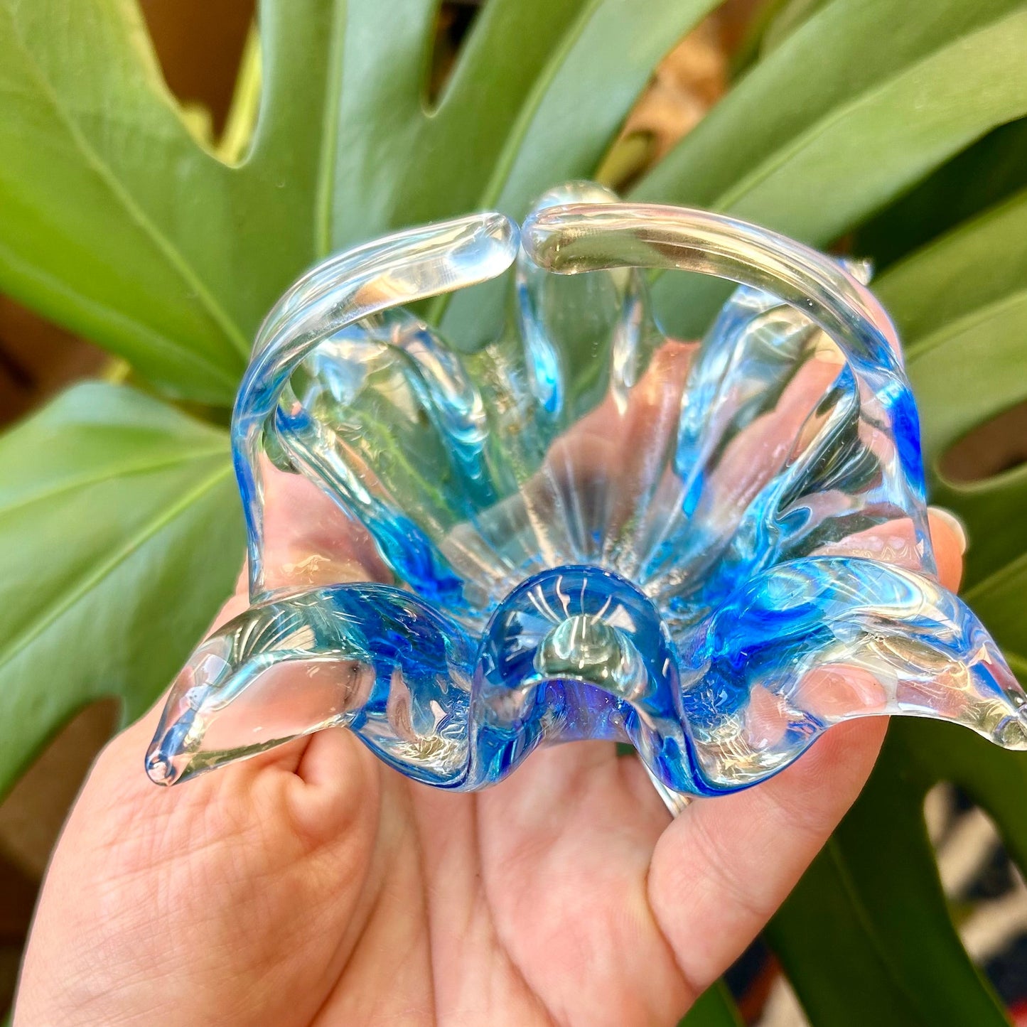 Hand holding a blue glass shell-shaped object with green foliage in the background