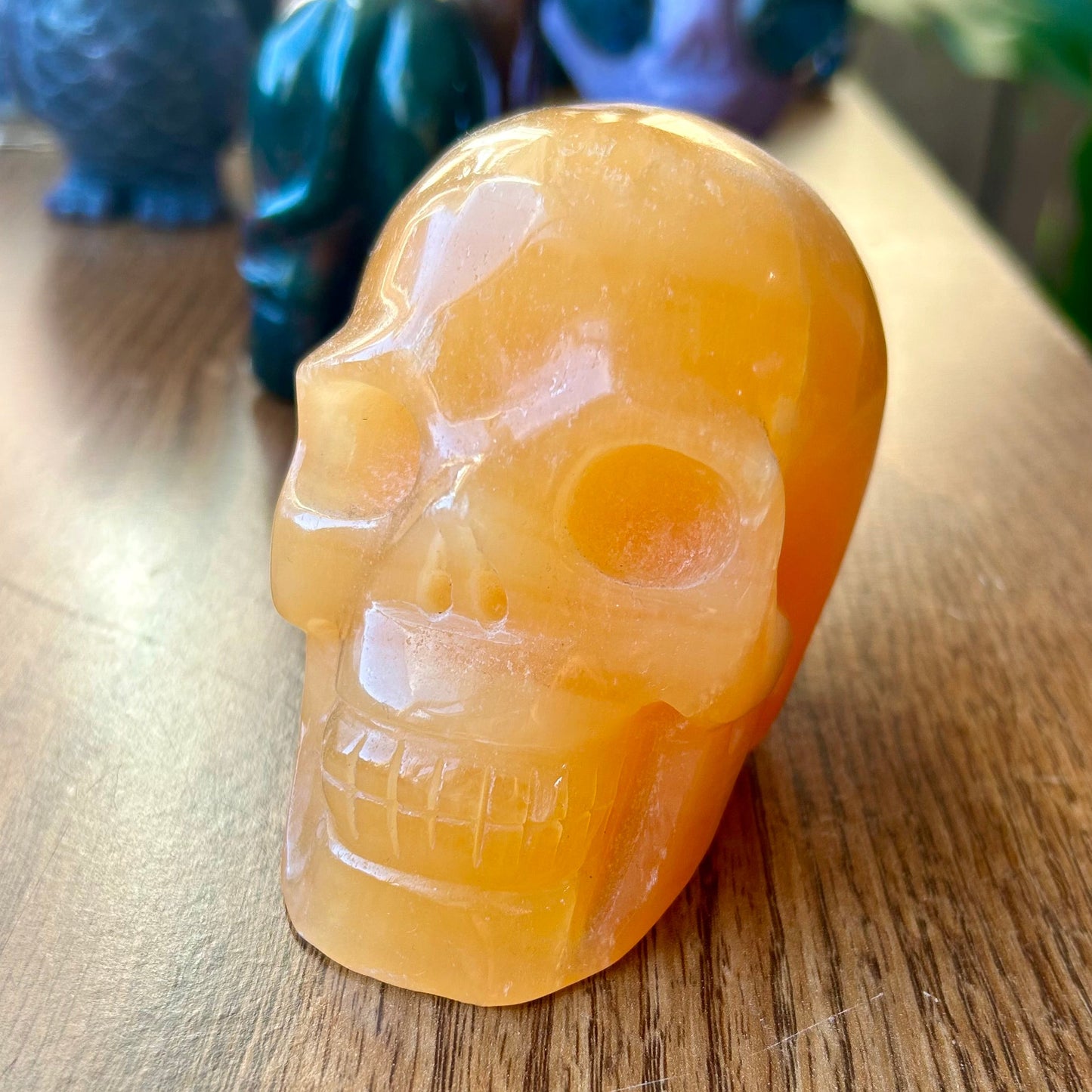 Orange crystal skull on a wooden surface with blurred colorful owl sculptures in the background.