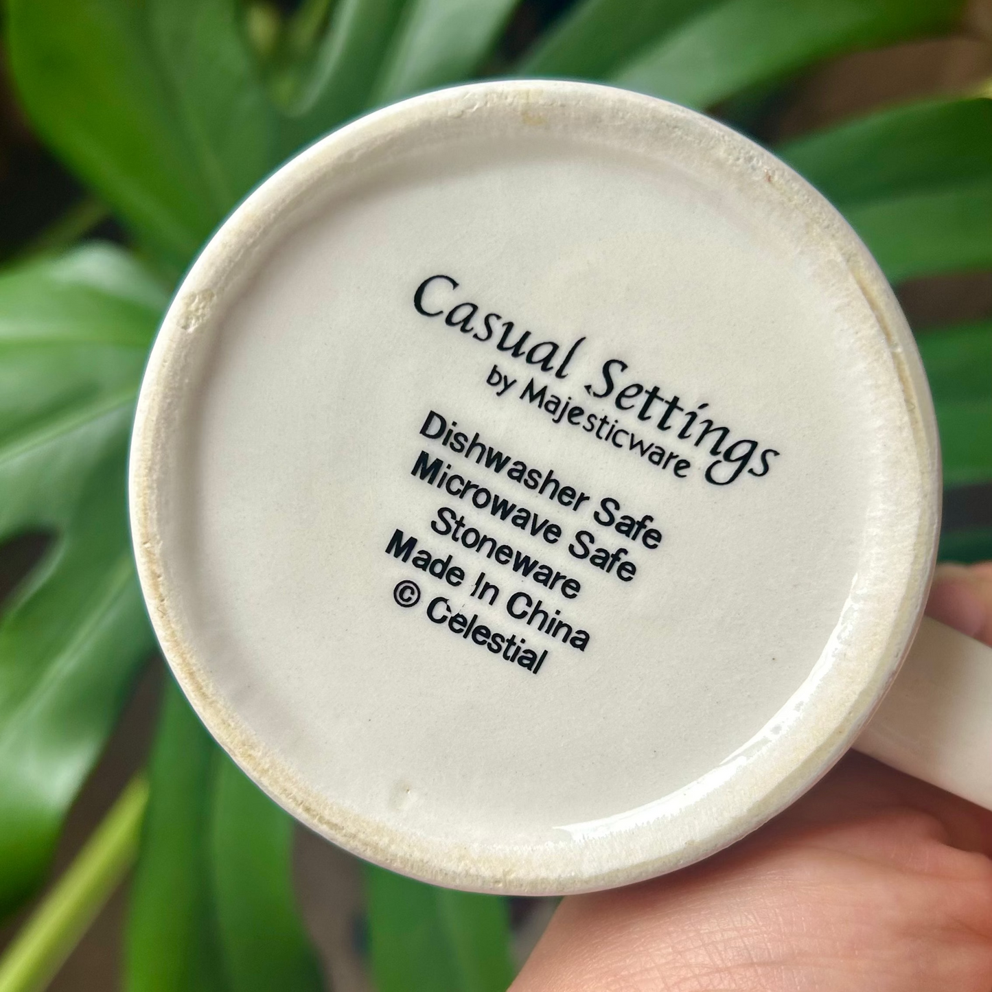 Close-up of a white mug with 'Casual Settings' text, held against a green leafy background.