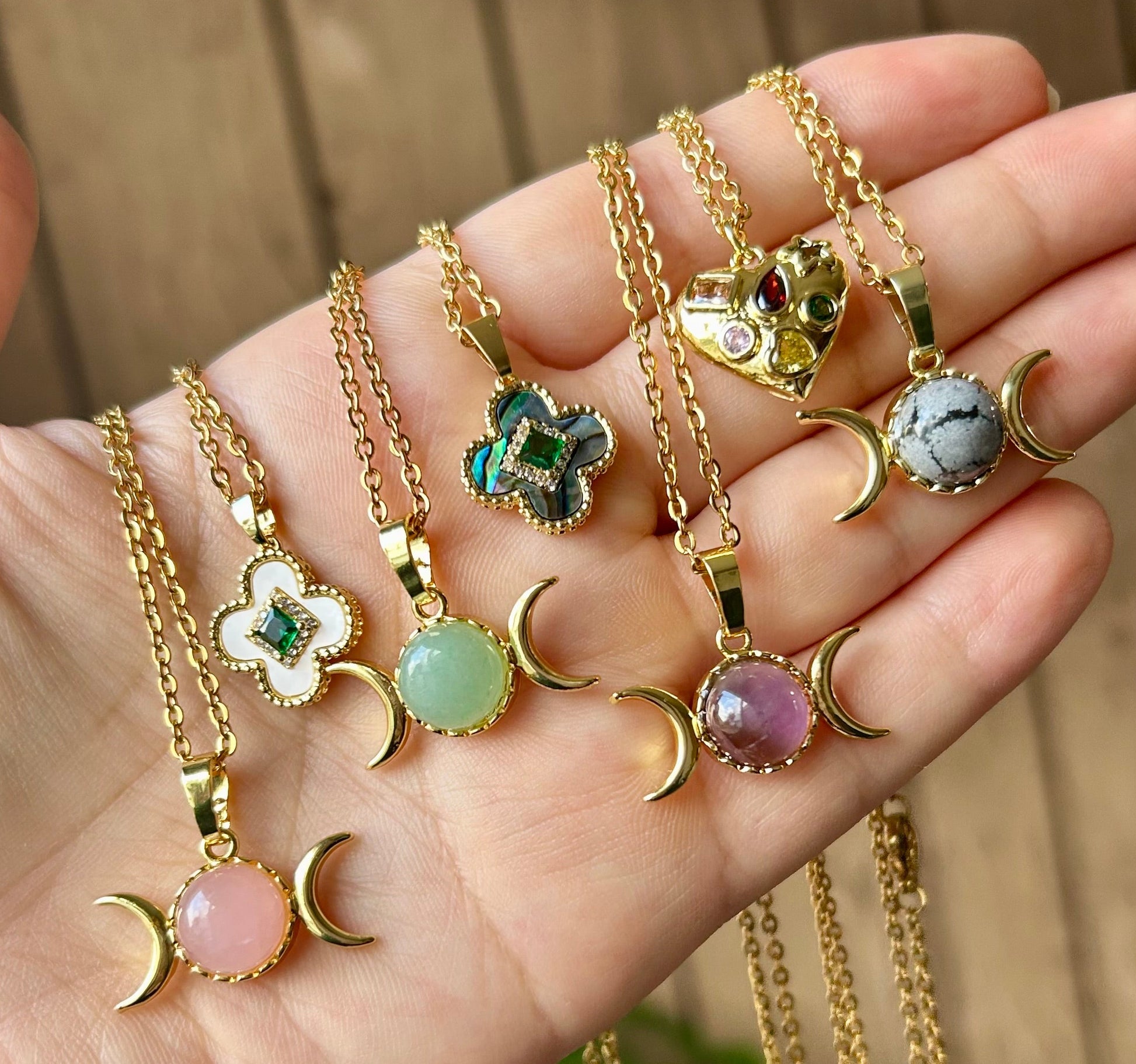 Hand holding multiple gold necklaces with various pendants against a blurred natural background