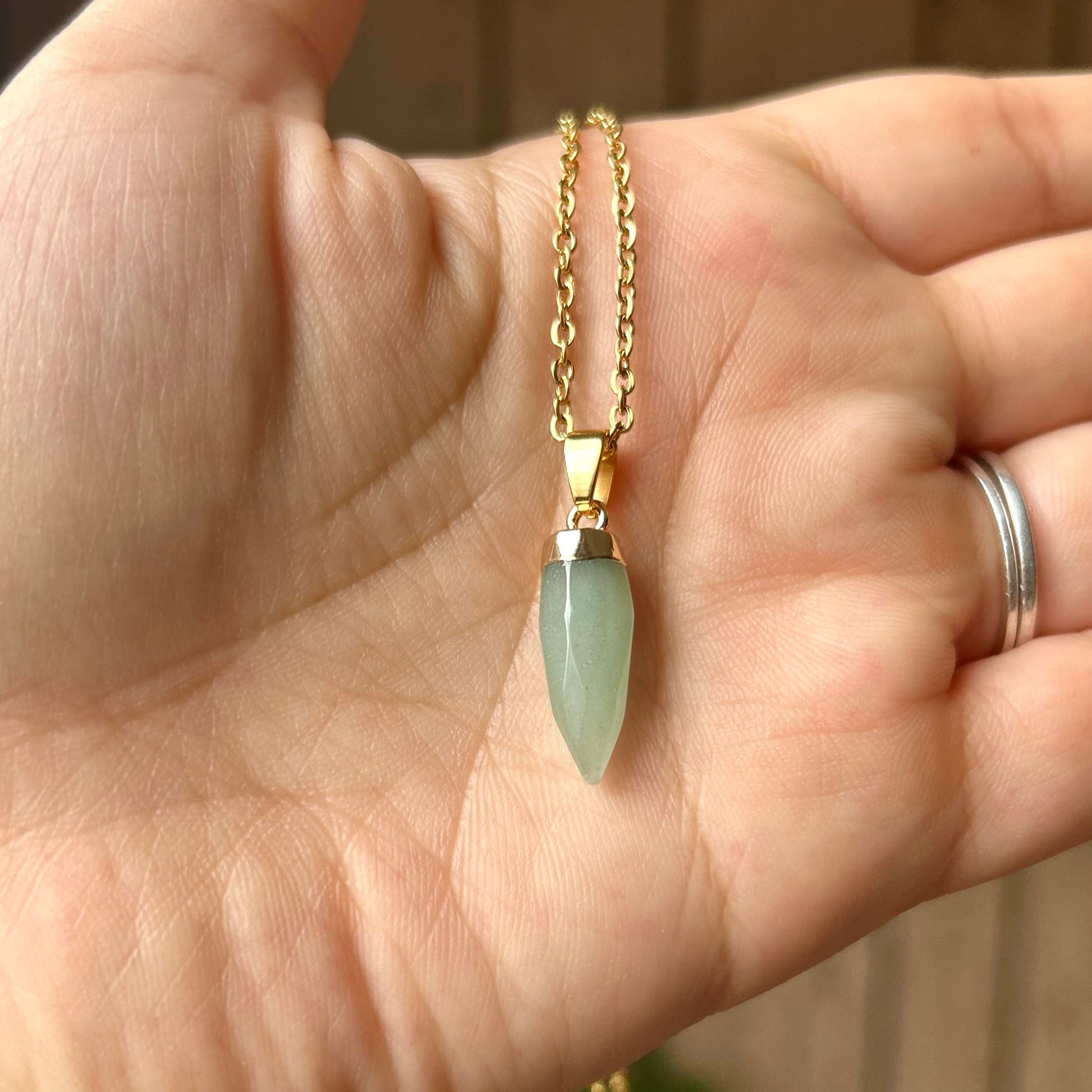 Hand wearing a gold necklace with a green pendant against a blurred background