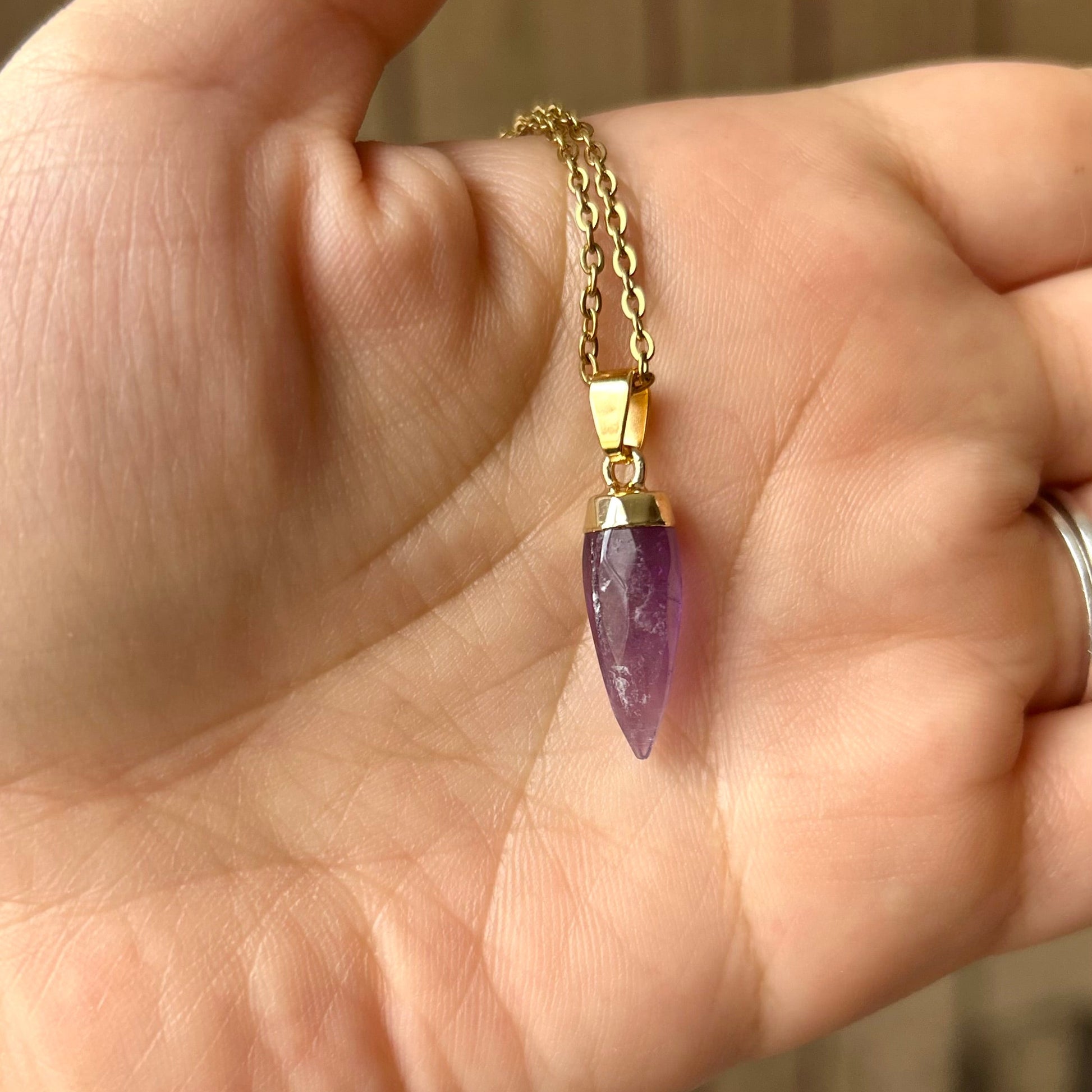 Hand wearing a gold bracelet with a purple gemstone pendant against a blurred background