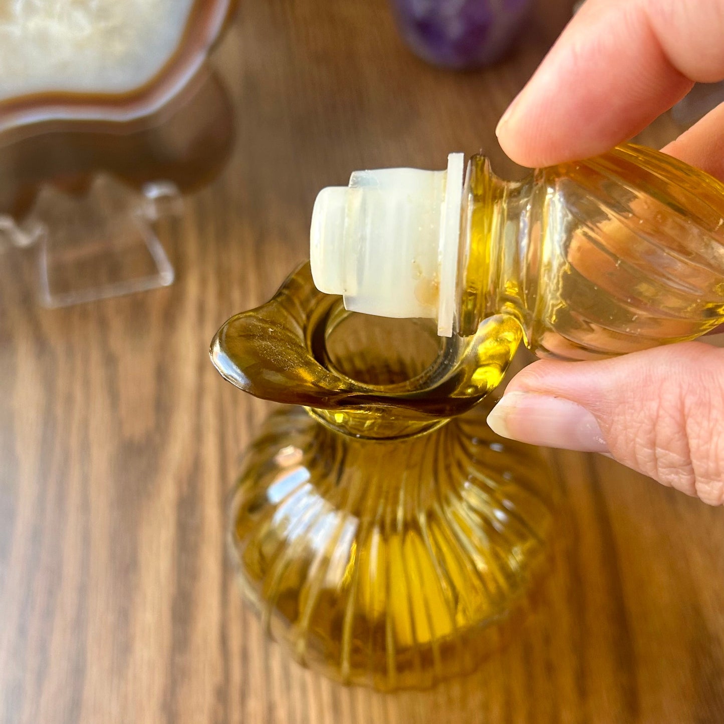 Hand pouring oil into a glass bottle on a wooden surface with various items in the background.