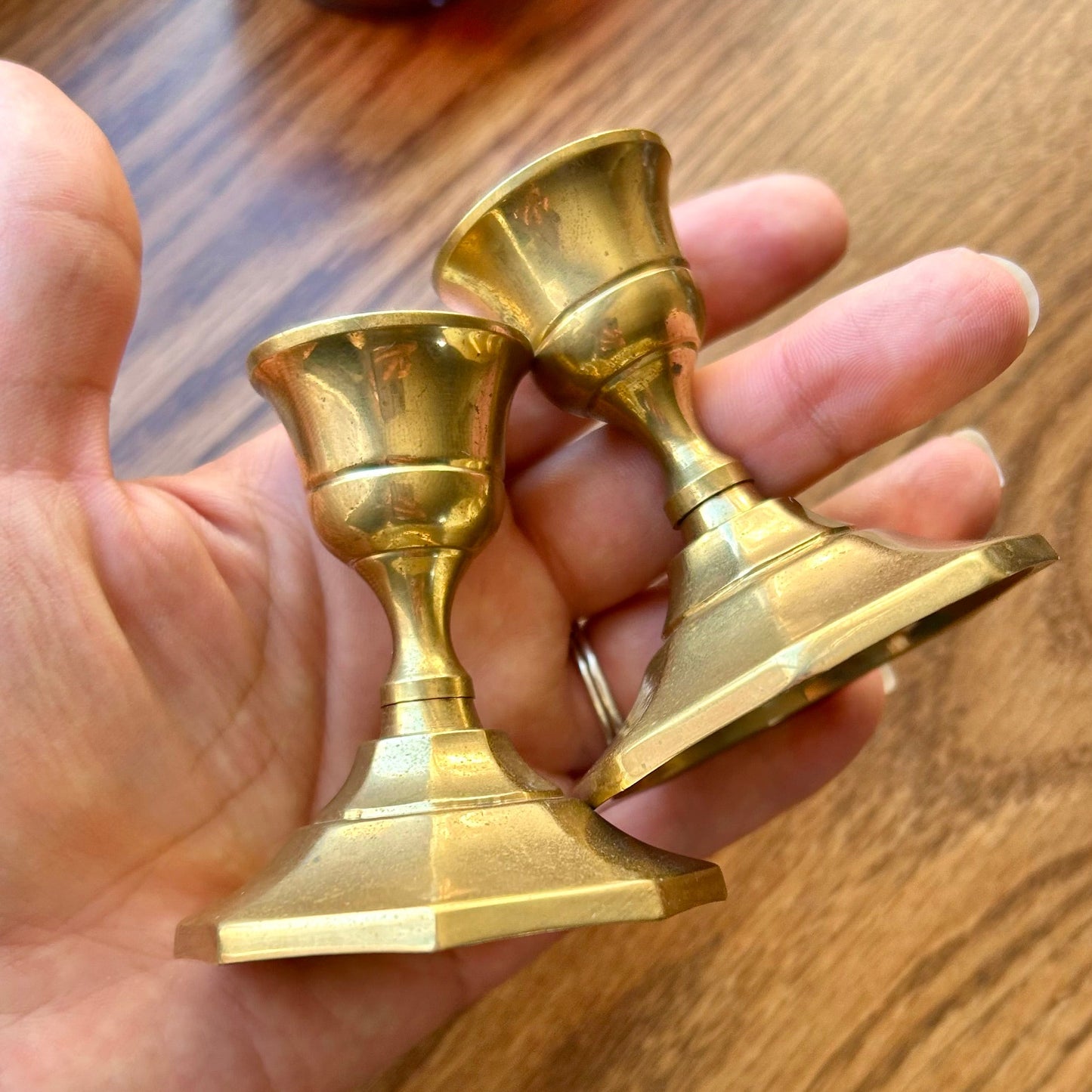 Hand holding two small gold candle holders on a wooden surface