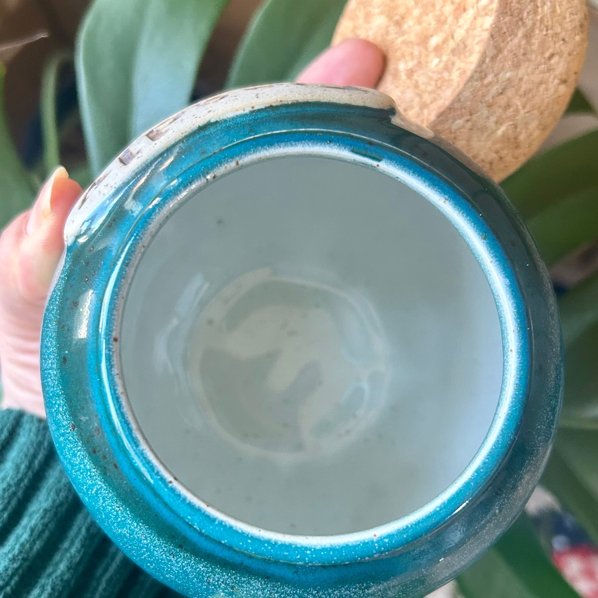 Close-up of a blue glass container with a cork stopper held by a hand, with green leaves in the background.
