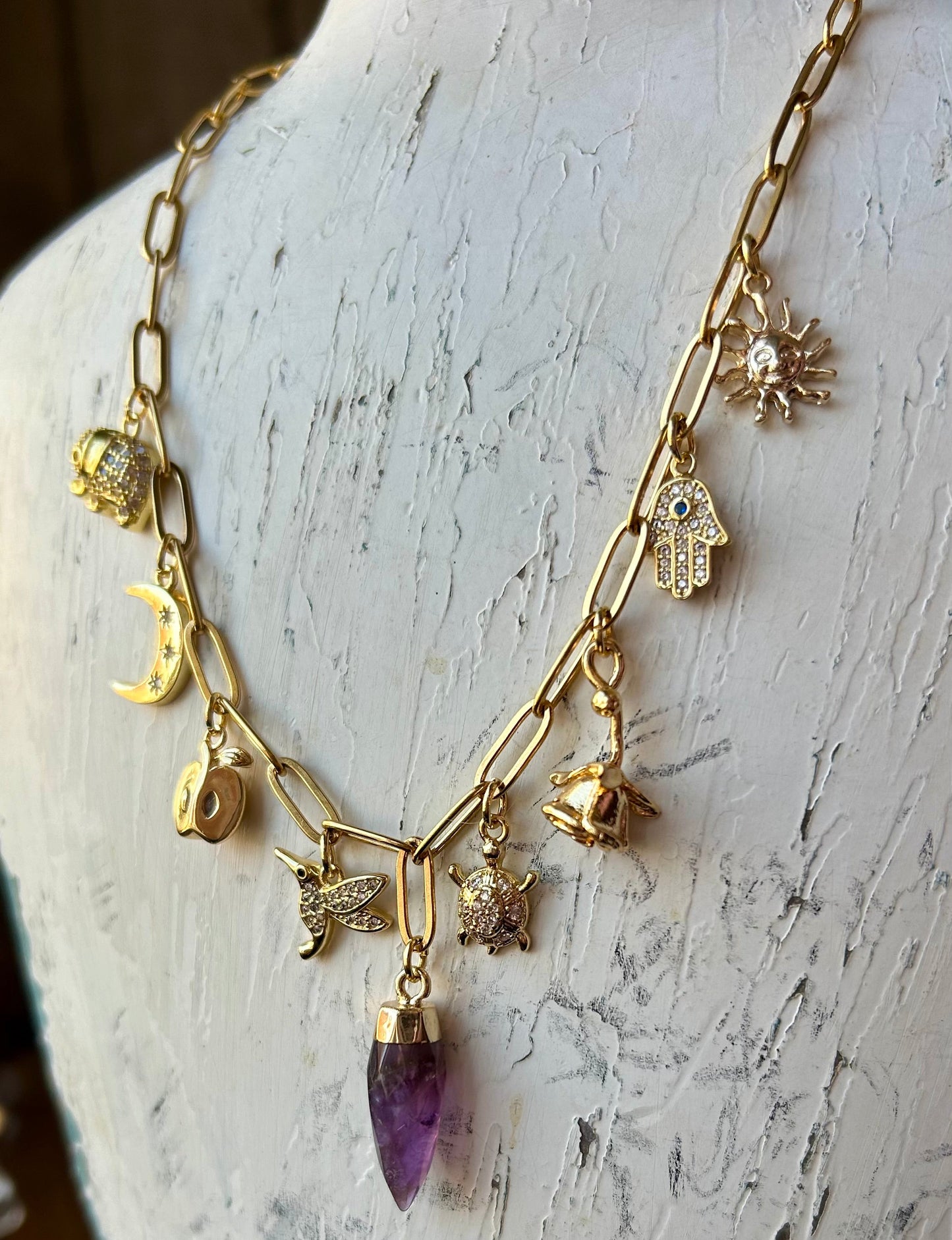 Gold necklace with various charms and a purple pendant on a white mannequin bust.