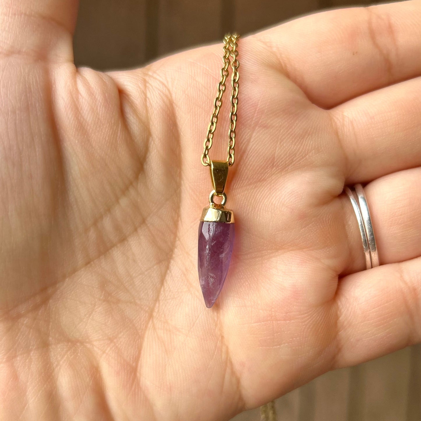 Hand holding a gold necklace with a purple pendant against a blurred background