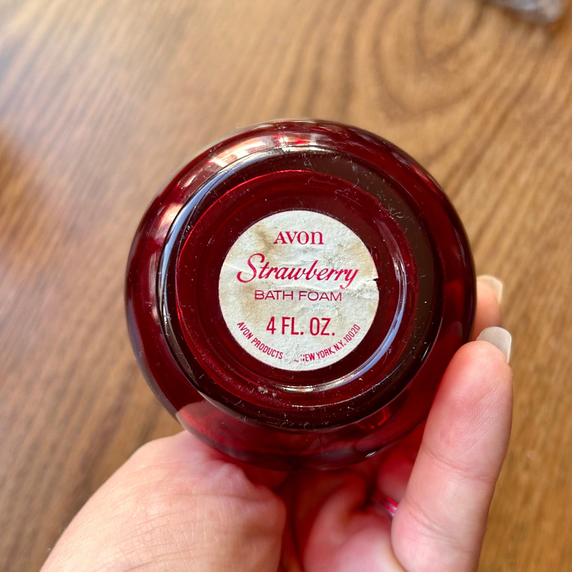 Hand holding a small red jar of Avon Strawberry Bath Powder on a wooden surface.