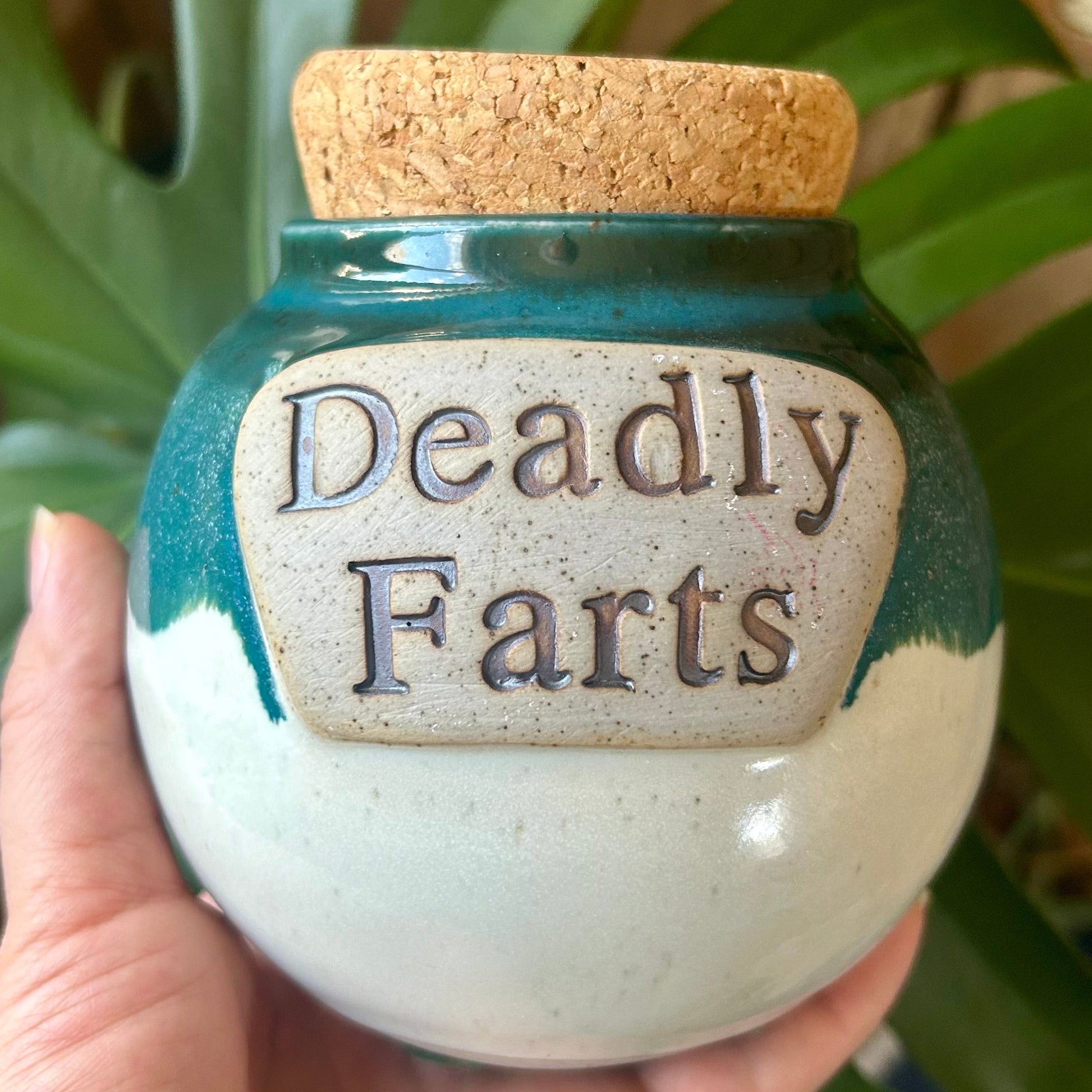 Jar labeled 'Deadly Farts' held in a hand with a green leafy background